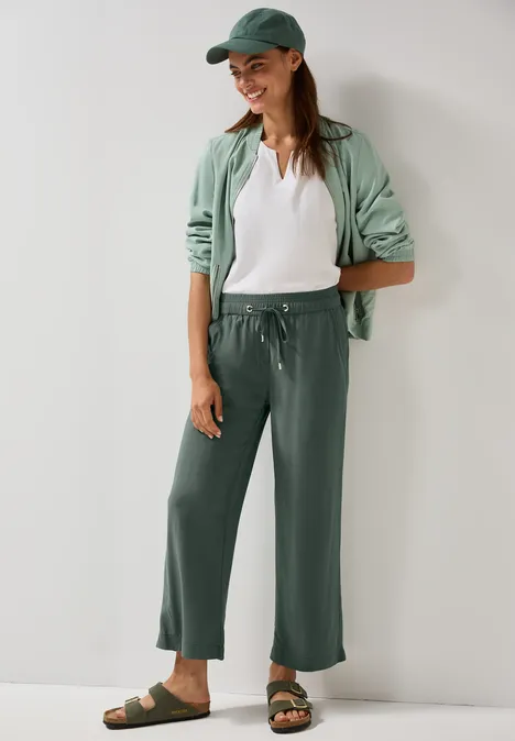 Frau in mintgrüner Kappe, Jacke und Hose, weißes Shirt, Sandalen, lässiger Stil.